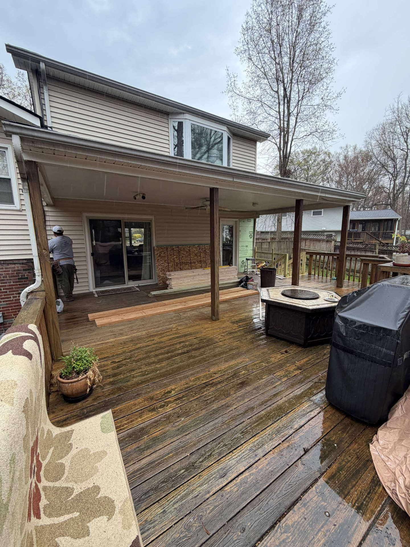 Before photo of the back of a two-story home with an open covered deck, exposed support posts, weathered decking, and a sliding glass door — taken on a rainy day before the addition was framed in.