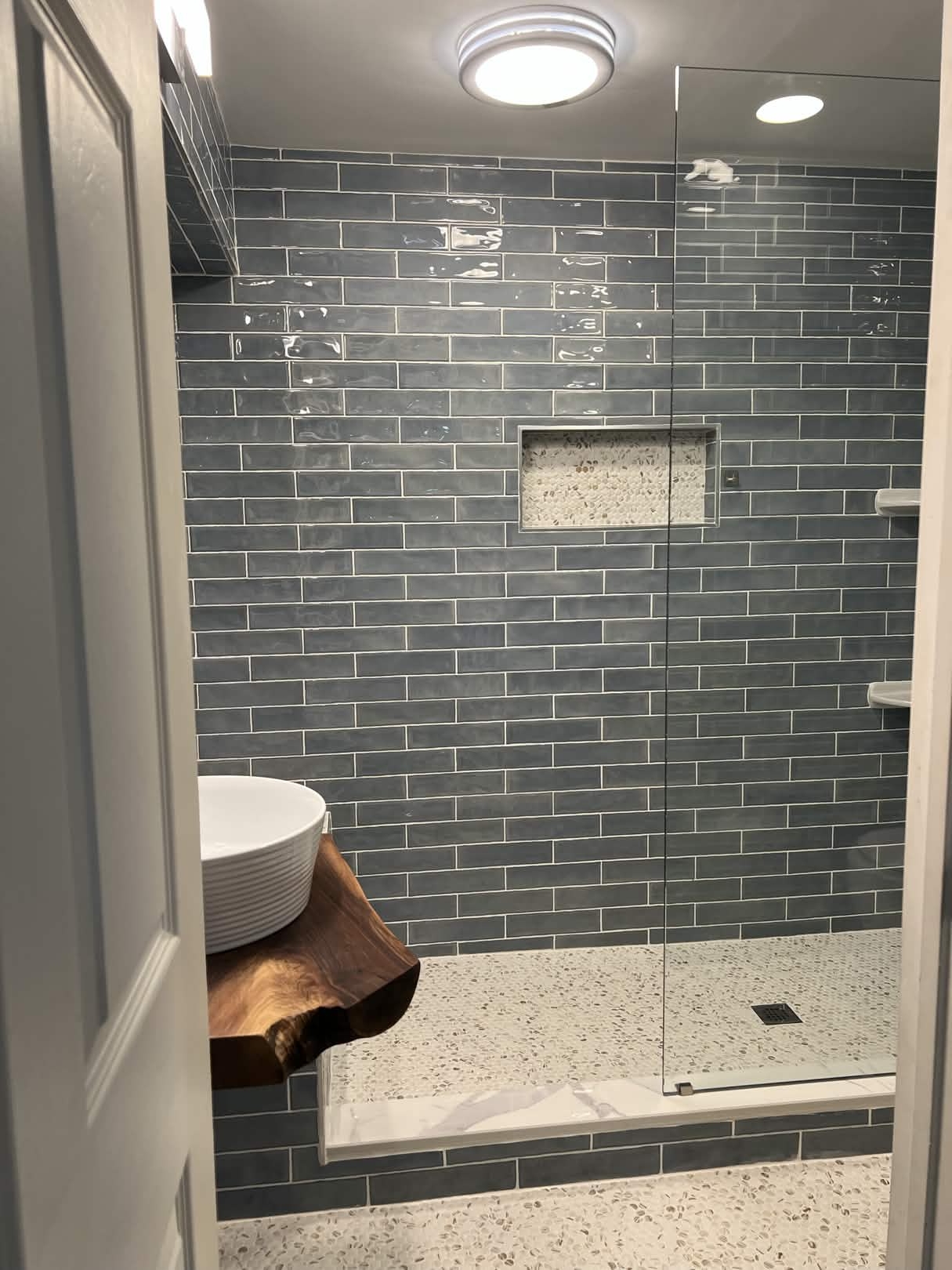 View through the bathroom doorway showing blue glass subway tile walls, a pebble mosaic floor, a tiled built-in shower bench with marble cap, a pebble-lined recessed shower niche, and the live-edge walnut vanity in the foreground.