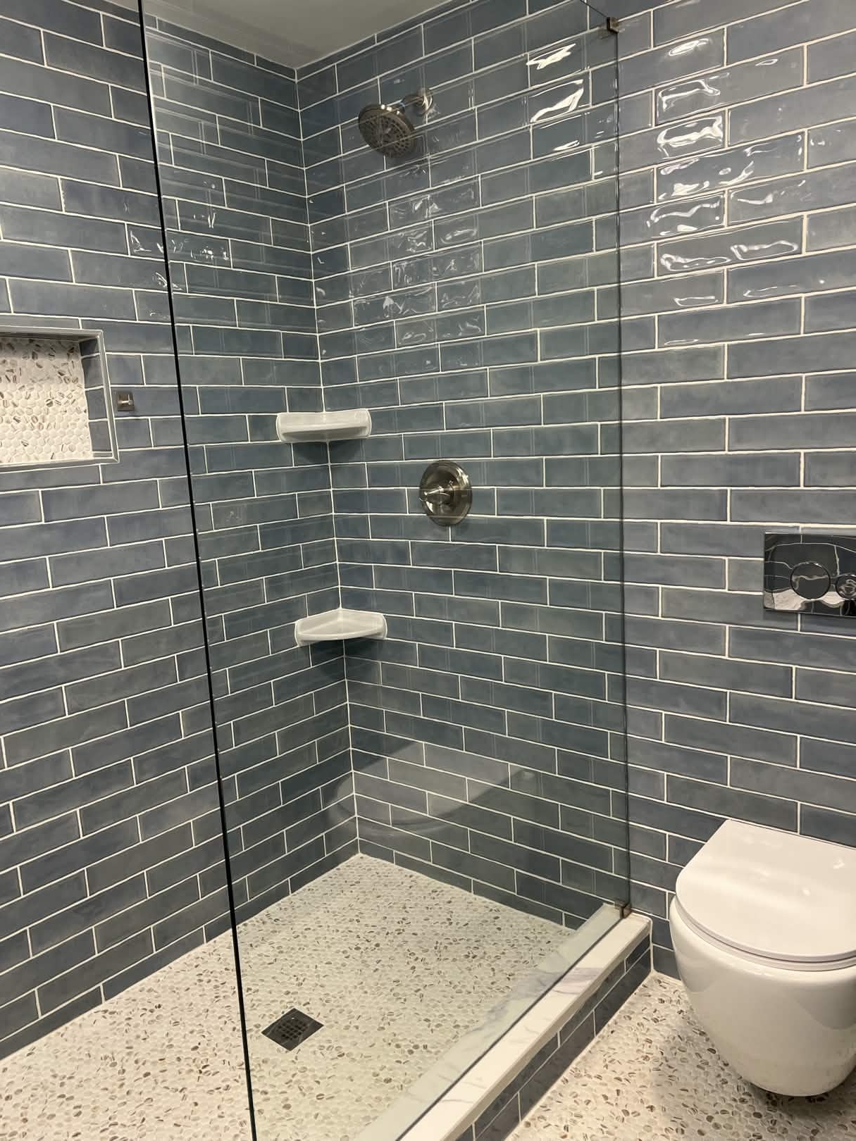 Custom walk-in shower with handmade blue glass subway tile, frameless glass panel, pebble mosaic shower pan, two white corner shelves, a pebble-lined recessed niche, and a wall-hung white toilet beside it.