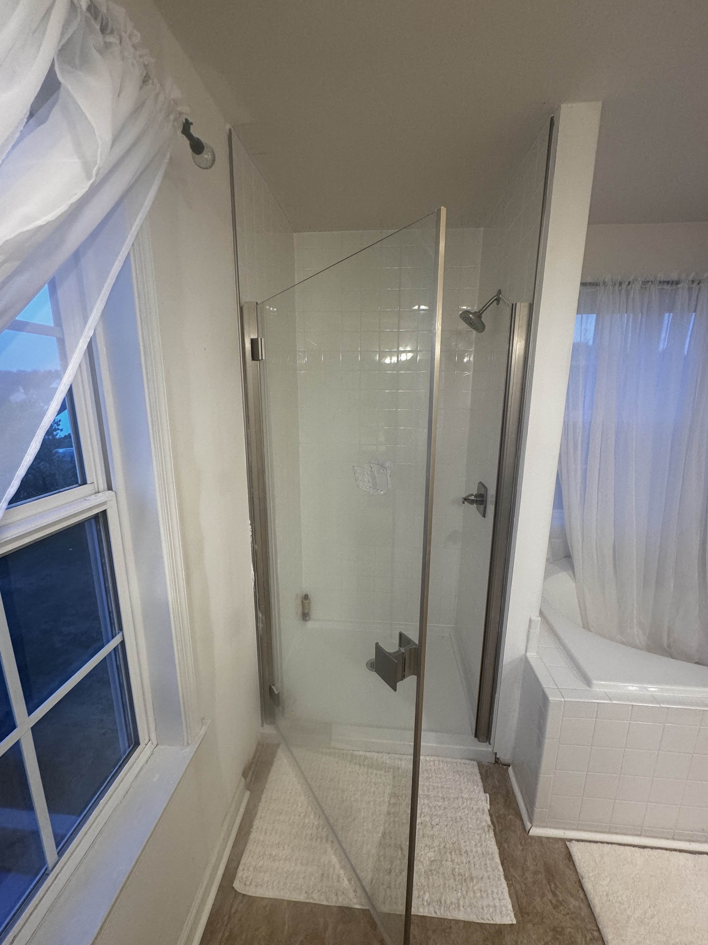 Compact shower stall finished with bright white square wall tile, a low-profile white shower pan, brushed-nickel showerhead and trim, and a custom frameless glass door and side panel between a window wall and a built-in tub deck.
