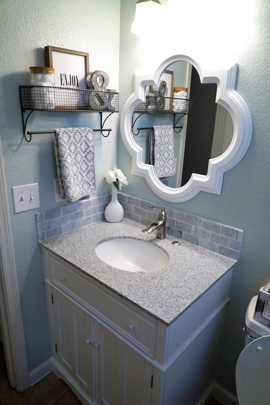 After photo of a remodeled coastal-style powder room with soft seafoam walls, white shaker vanity, speckled granite countertop with undermount sink, marble subway tile backsplash, and a decorative white quatrefoil mirror.