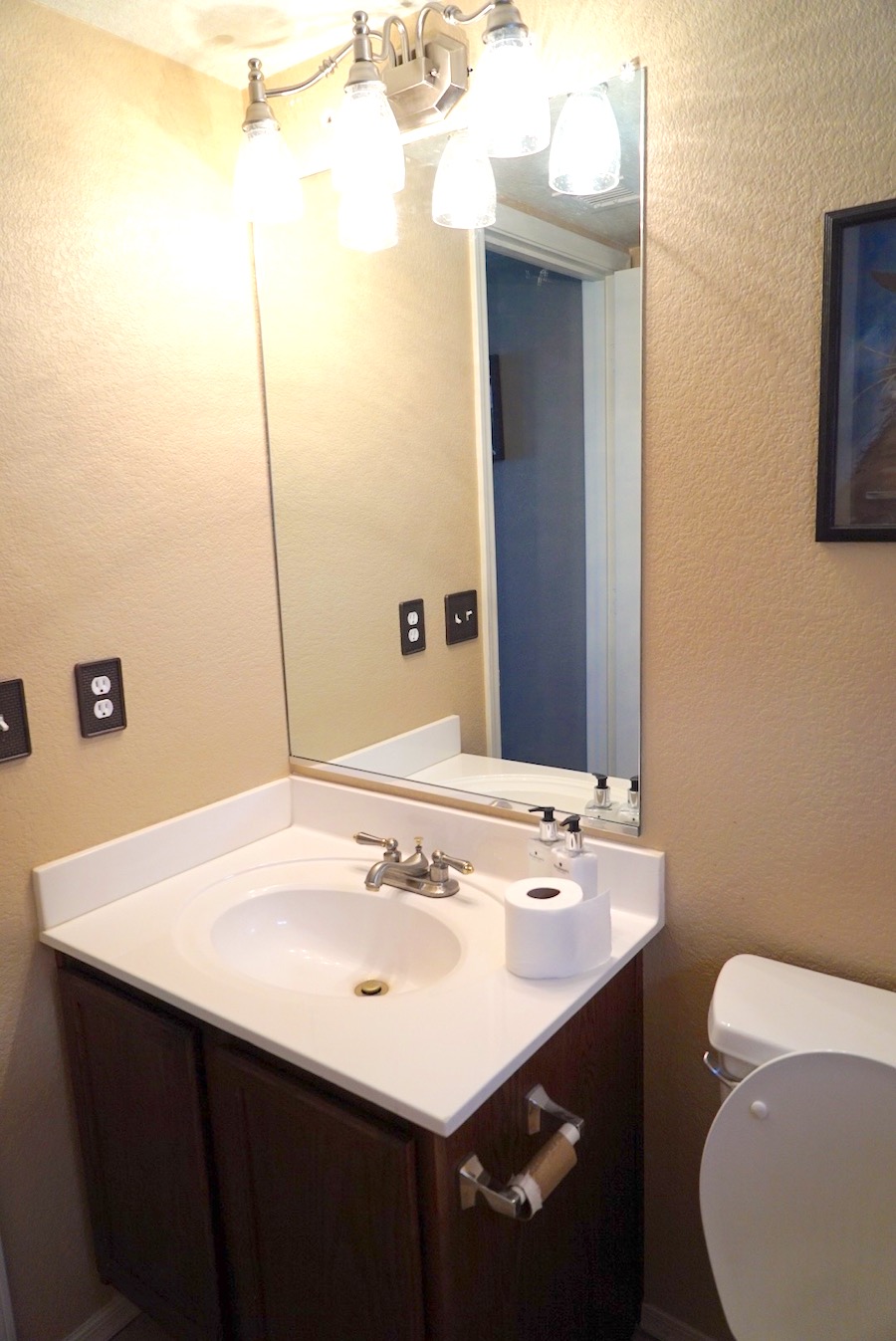 Before photo of a dated powder room with beige textured walls, dark builder-grade vanity, white drop-in sink, large frameless mirror, and brushed-nickel light bar.