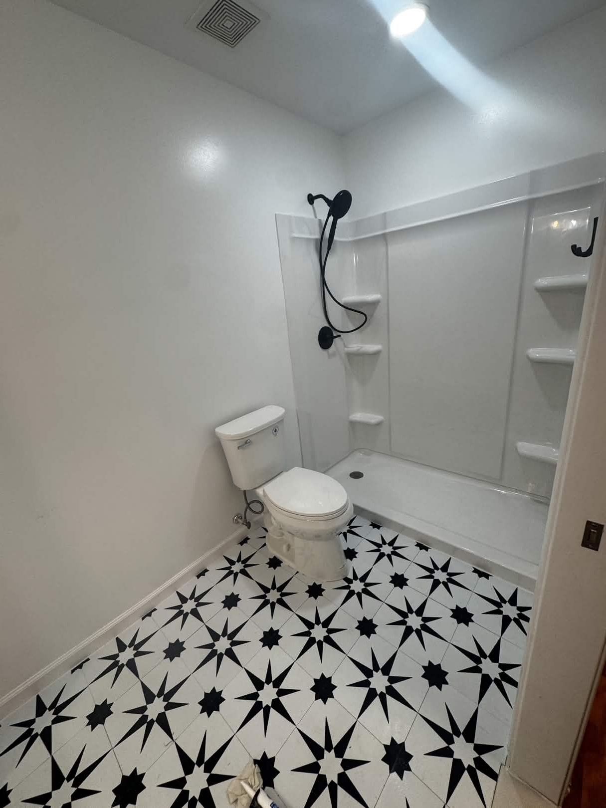 Newly finished full bathroom with a black-and-white star-pattern porcelain tile floor, a white one-piece fiberglass shower surround with built-in corner shelves, matte-black showerhead and trim, and a white comfort-height toilet.