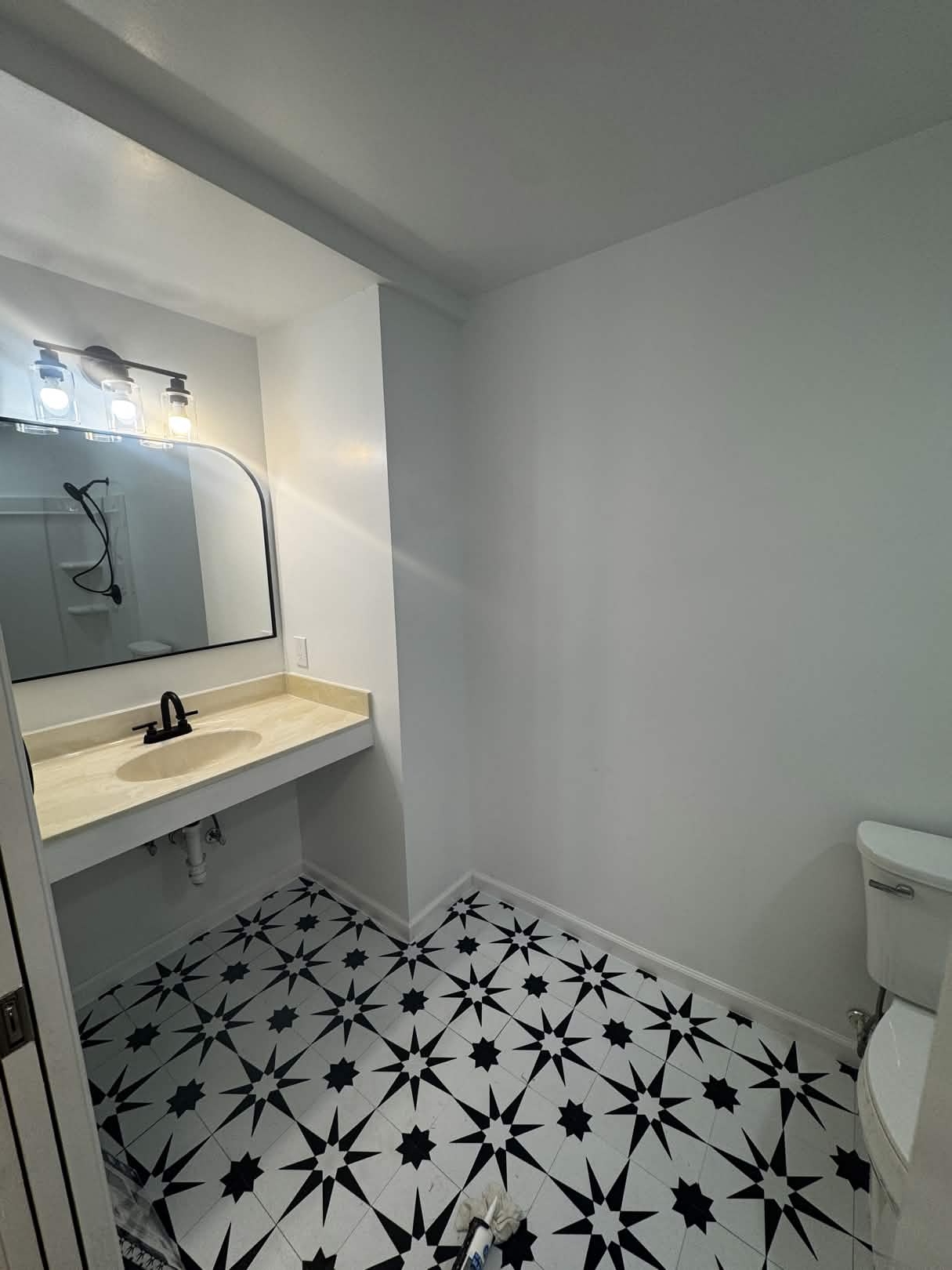 Built-in vanity nook with a cream cultured-marble top, white wall-hung sink, matte-black faucet, arched black-framed mirror, and a three-light matte-black vanity fixture above a black-and-white star-pattern tile floor.