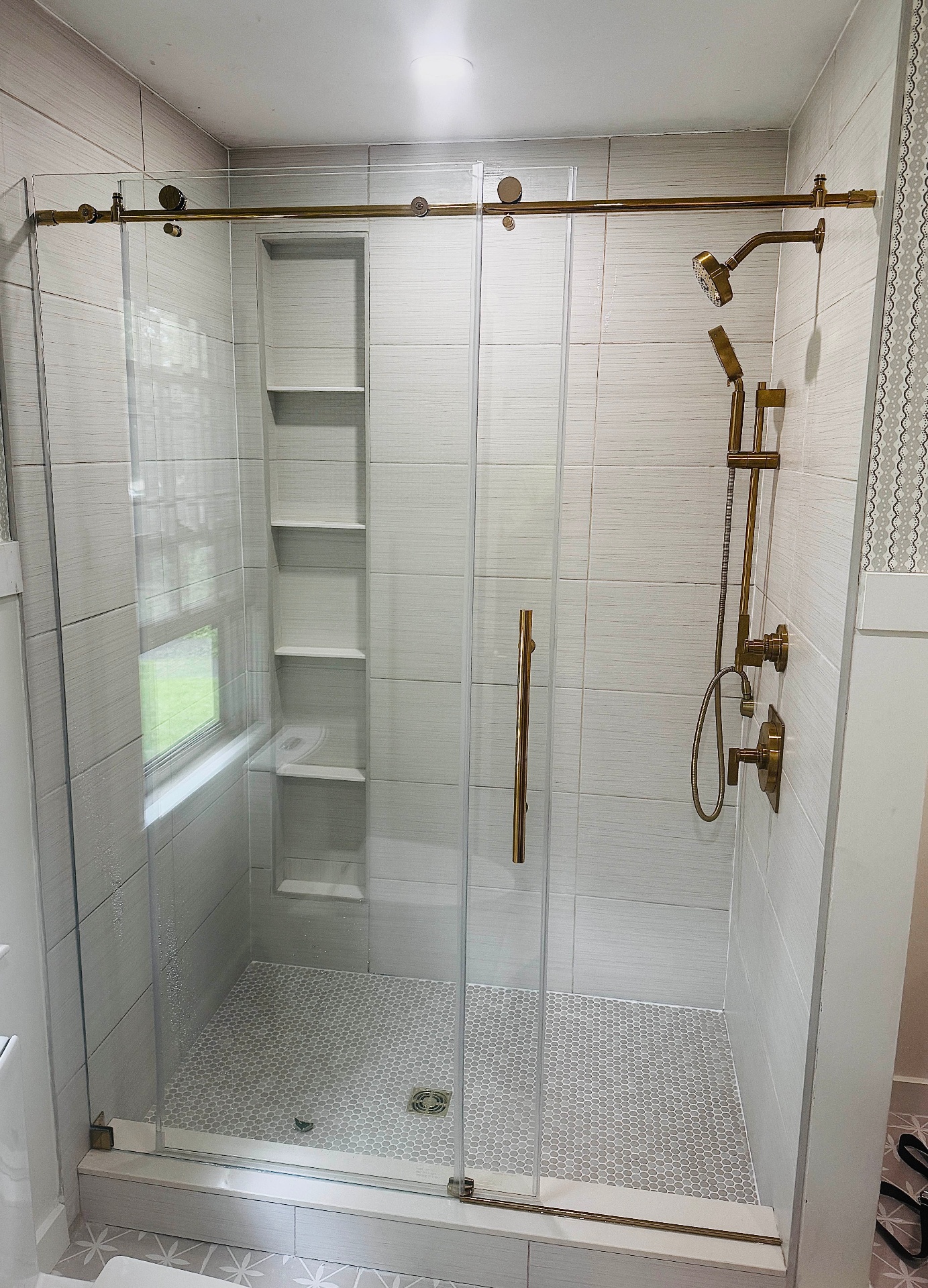 Finished walk-in shower with large-format light grey tile, tall built-in shelving niche, white hexagon mosaic shower pan, frameless sliding glass doors, and brushed-gold rainhead, handheld sprayer, and shower trim.