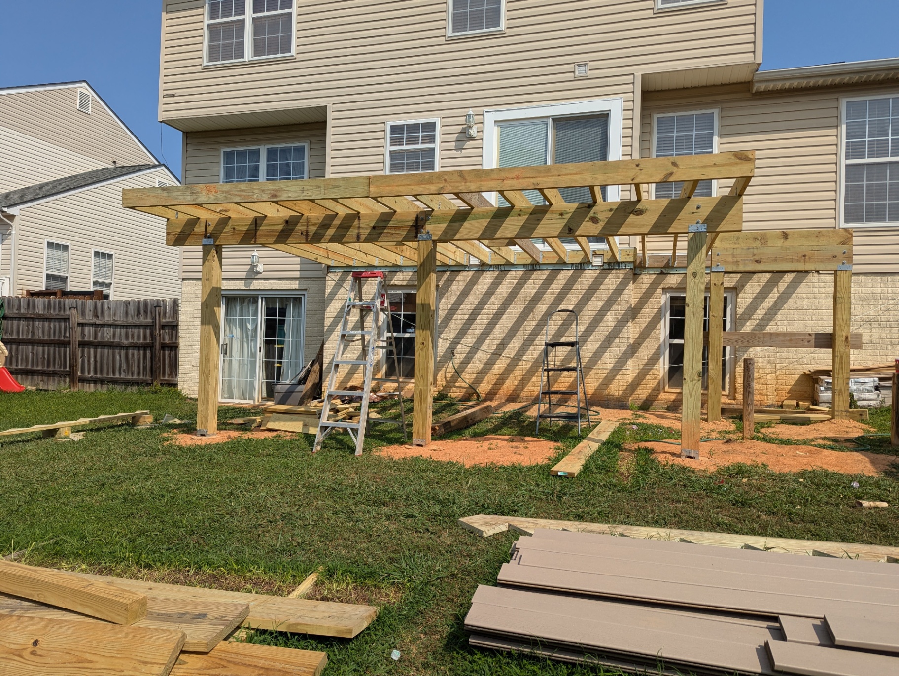 Pressure-treated wood pergola-style frame with support posts, beams, and joists set against the back of a two-story home during the early framing stage of a custom deck build.