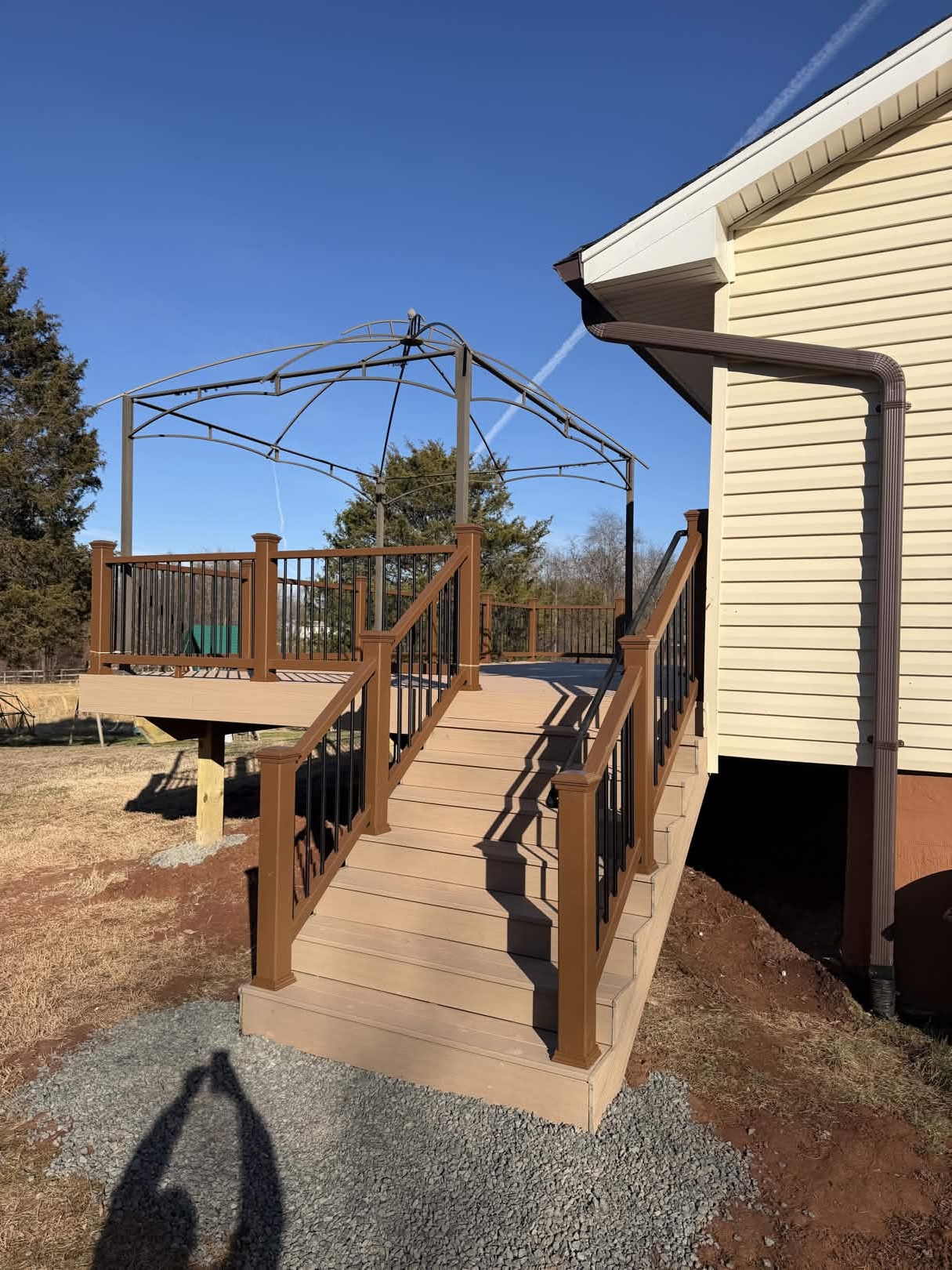 Wide composite back-deck staircase with brown railing posts, black aluminum balusters, and tan stair treads landing on a fresh gravel pad.