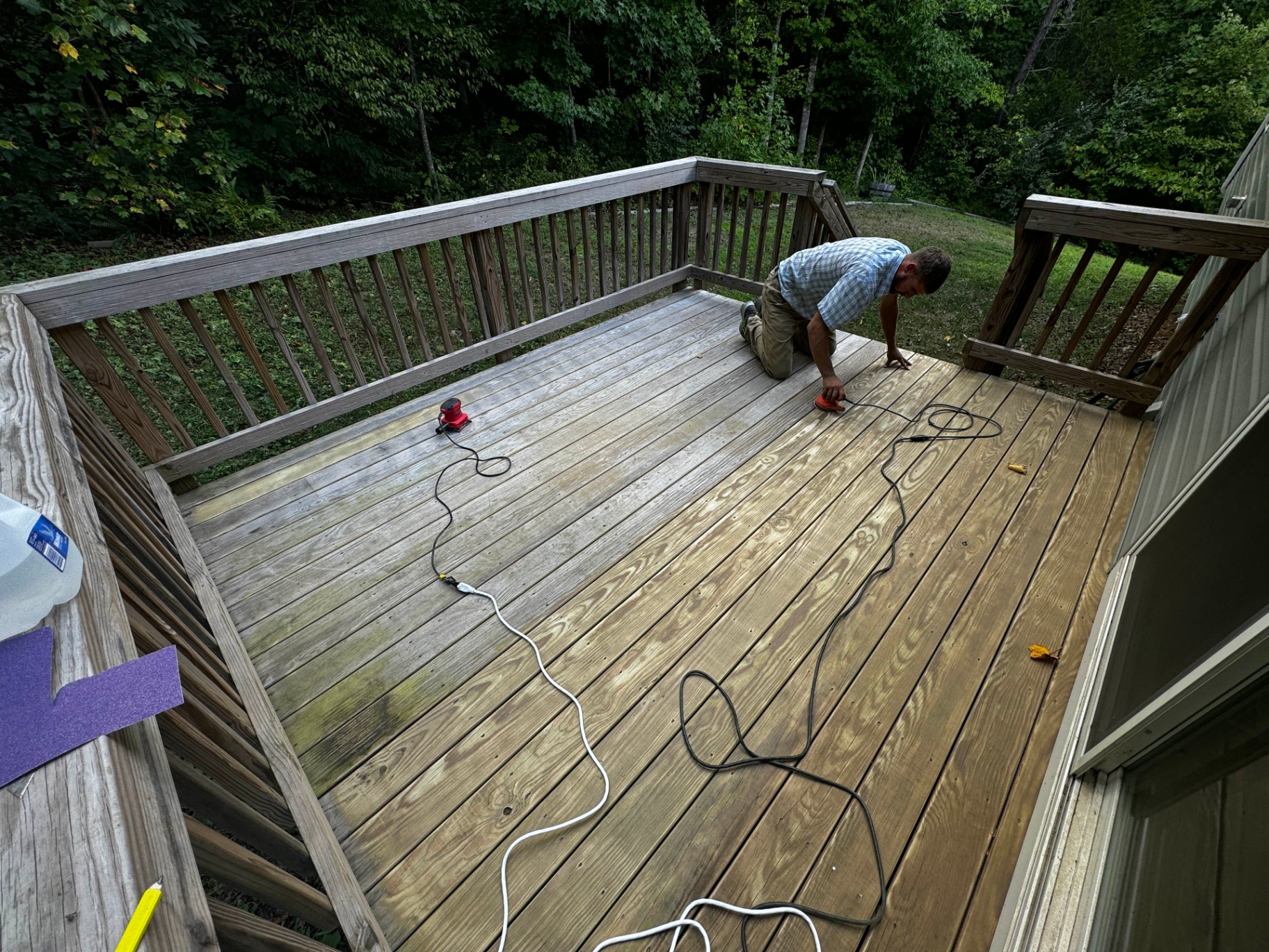 Carpenter sanding a weathered, gray pressure-treated wood deck with an orbital sander, prepping the boards and railings for a fresh stain.