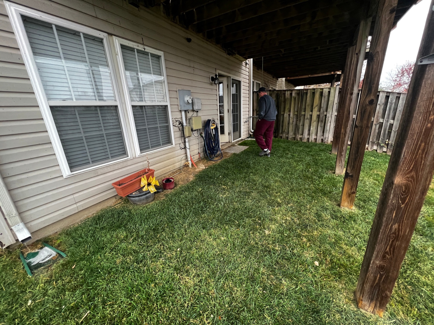 Before photo of a narrow grassy backyard area beneath an existing wood upper deck, with utility meters on the home's siding and a wood privacy fence along the property line.