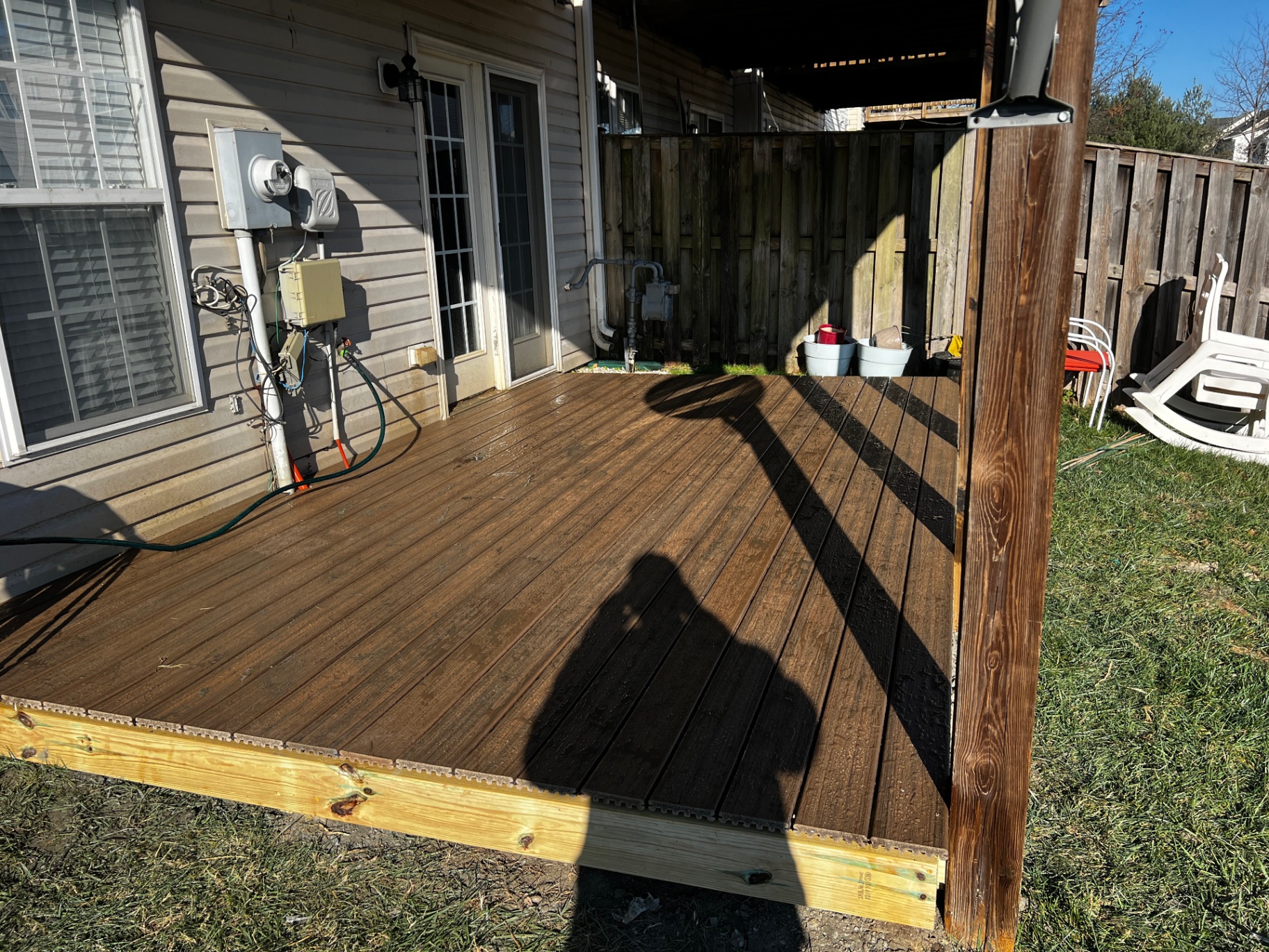 Finished ground-level composite deck with rich brown grooved decking boards installed beneath an existing upper deck at the back of a residential home, with a wood privacy fence and lawn surrounding it.
