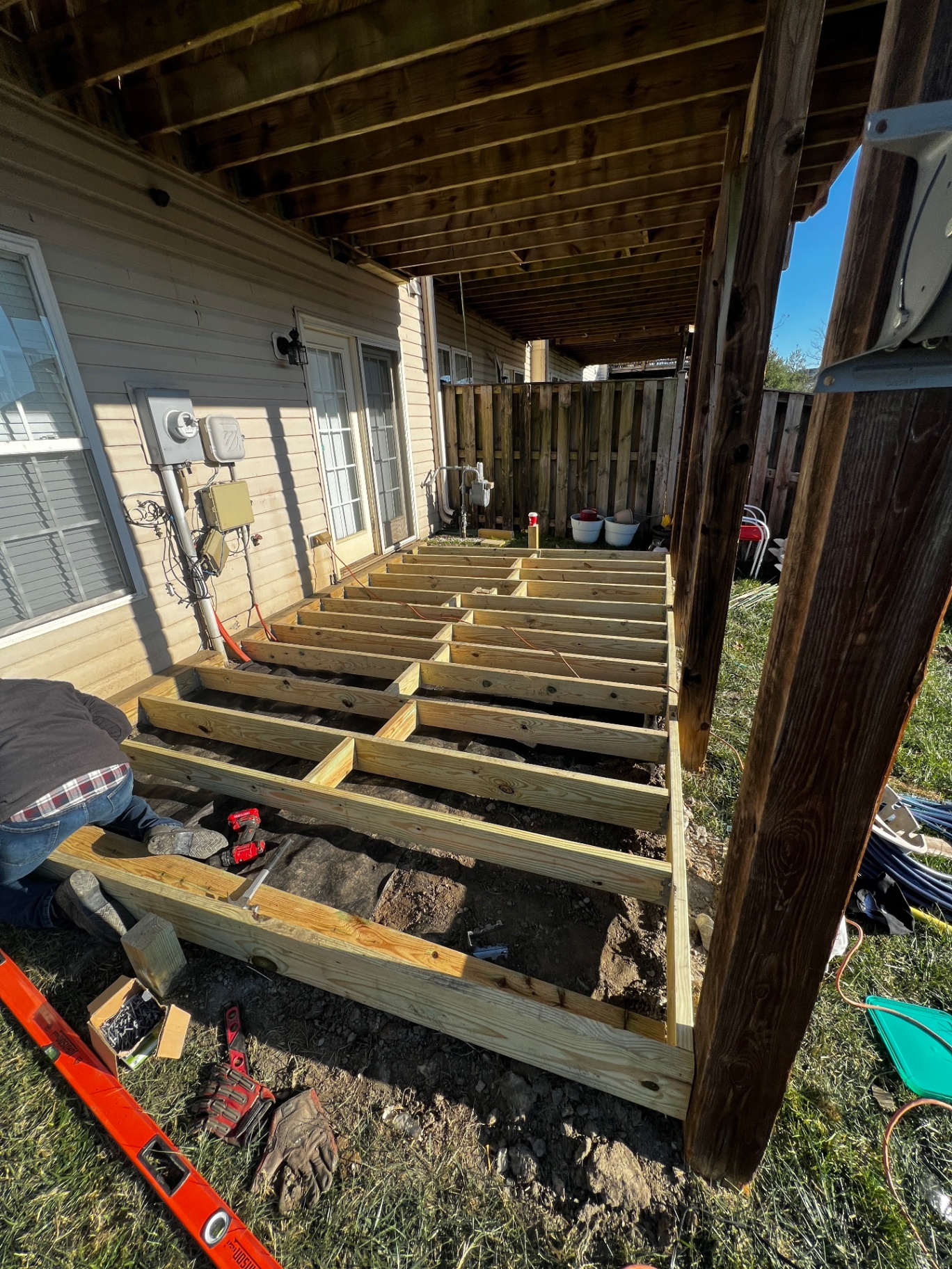 Pressure-treated floor joist framing for a new ground-level deck, set on rim joists between a residential home and a privacy fence with tools and a level on the ground.