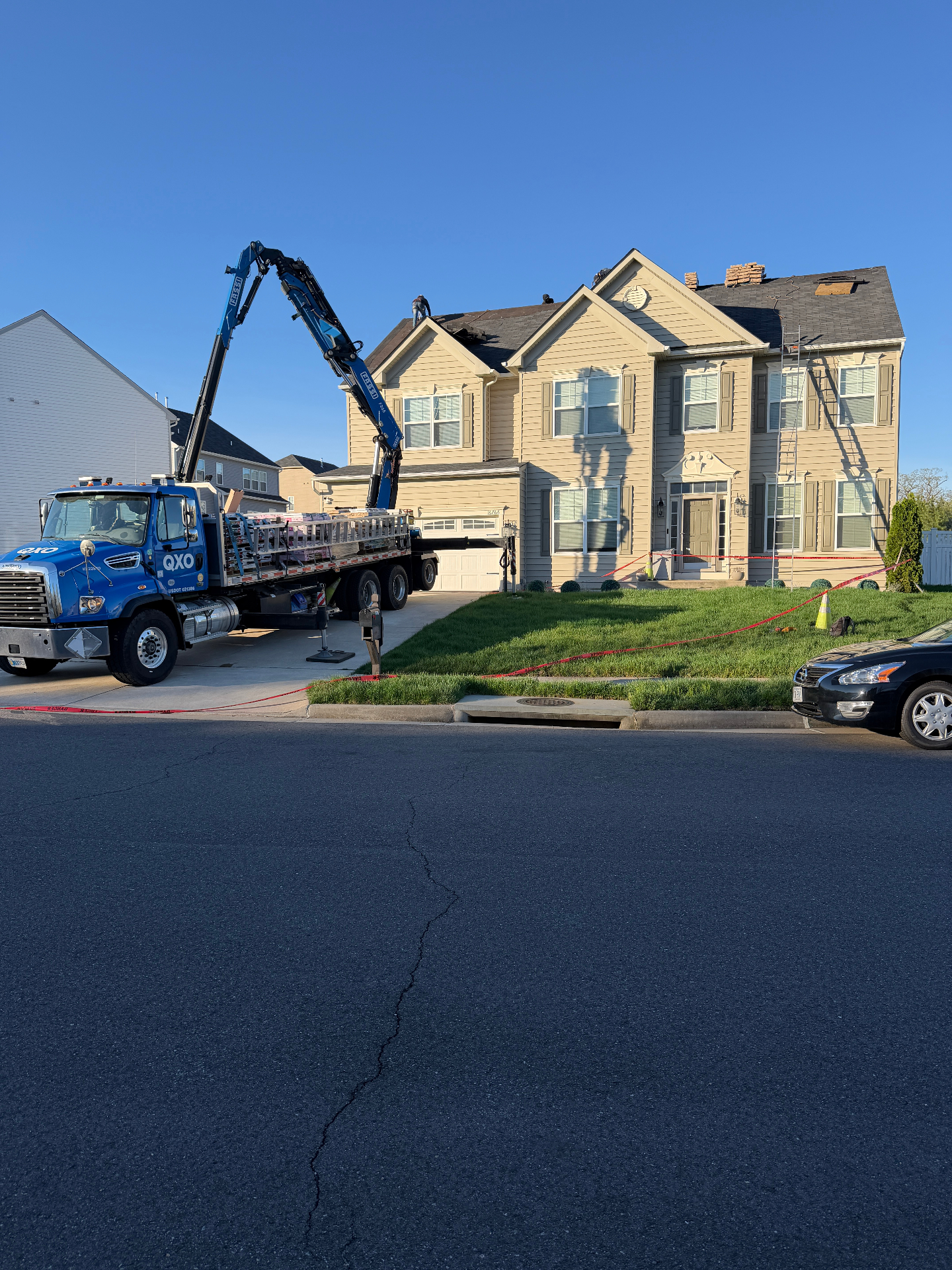 QXO blue boom-truck parked at the curb of a two-story residential home, lifting bundles of asphalt shingles onto the roof for a full roof replacement by Perpetual Improvements.