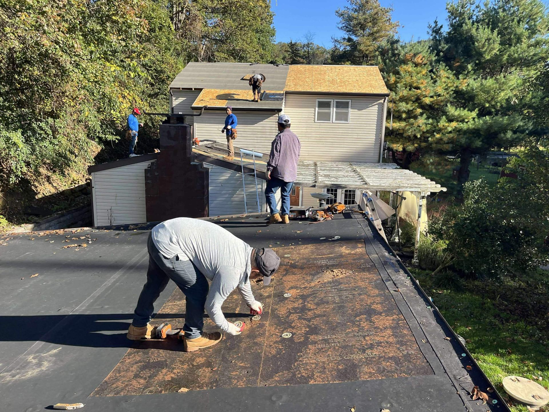 Perpetual Improvements roofer installing new EPDM rubber membrane on a residential flat roof while additional crew members work on the adjacent sloped shingle roof of the main house.